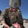 A U.S. Marine in full combat gear and MARPAT camouflage is providing tactical combat casualty care (TCCC) to a downed fellow Marine. The casualty has simulated facial wounds consistent with a training exercise, including moulage (simulated blood and injuries) applied to the face. The setting appears to be a military training environment based on visible structures and terrain in the background.