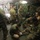 The image shows military personnel inside what appears to be a field medical tent or aid station. Several individuals in military uniforms are attending to what appears to be a casualty or patient on the ground, with medical treatment being administered to a lower extremity. The setting is dimly lit with motion blur on some figures, suggesting active and urgent movement.