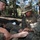 Two U.S. Marines in MARPAT digital camouflage uniforms and tactical vests are seated on the ground in a forested area. One Marine is attending to the bare foot of another person, appearing to provide first aid or blister care. The setting suggests a field training exercise in a mountainous or high-elevation forest environment.