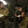 The image shows what appears to be a military field medical station inside a camouflage-patterned tent. Three individuals in military attire are present, with one seated patient receiving what appears to be a medical examination or treatment near the face, while another prepares medical supplies. The setting includes medical storage equipment and portable lighting.
