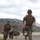 Military personnel in full combat gear are running across a paved road during what appears to be a field training exercise. Multiple soldiers wearing MARPAT (Marine pattern) camouflage uniforms, plate carriers, and helmets are visible, with some taking cover in the background. The setting is a mountainous, arid terrain under overcast skies.