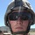 A military service member wearing MARPAT camouflage uniform and combat helmet is shown in close-up portrait. The individual has red splatter marks on their face and neck, consistent with training exercise simulated casualty makeup or paint marking rounds. A military HMMWV (Humvee) vehicle is visible in the background on what appears to be a training range or road.