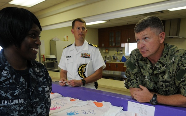 Three U.S. Navy personnel are gathered around a counter displaying handprint-decorated t-shirts reading 'CFAC Preschool 2010'. A female sailor in Navy Working Uniform (NWU) with the name tape 'CALLOWAY' is conversing with a senior officer in camouflage utilities, while a third officer in dress whites stands in the background. The setting appears to be a childcare or preschool facility, suggested by the Elmo decoration visible on the left wall.