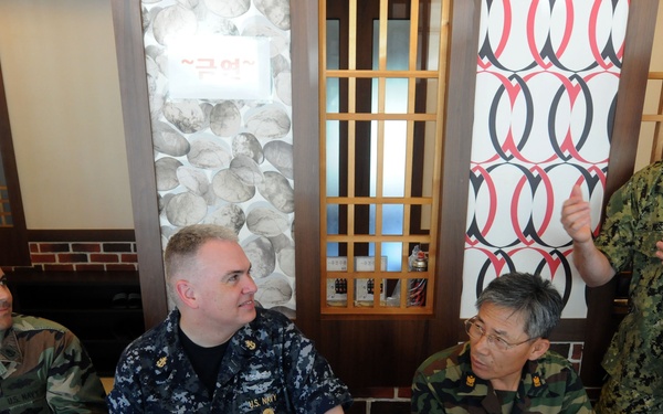 Four military personnel are seated and standing around a dining table in what appears to be a Korean restaurant, sharing a meal together. Three individuals wear different camouflage uniforms including U.S. Navy NWU and woodland patterns, while one appears to be a Korean military officer. The standing U.S. Navy officer appears to be speaking or making a point to the group.