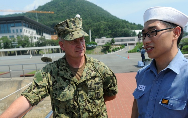 Two military personnel are standing on an outdoor terrace or walkway near a railing, appearing to be in conversation. One individual wears a US Navy digital camouflage uniform with a cover, while the other wears a light blue uniform with a white sailor's cap, consistent with an Asian naval service. The setting appears to be a military installation with buildings, green hills, and a construction crane visible in the background.