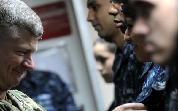 A senior military officer in U.S. Navy NWU Type II (desert camouflage) uniform is pinning or attaching something to the uniform of a sailor wearing NWU Type I (blue digital camouflage). The scene appears to be a military ceremony or recognition event taking place in an indoor shipboard or military facility corridor. Several other sailors in blue NWU uniforms are visible in the background observing.
