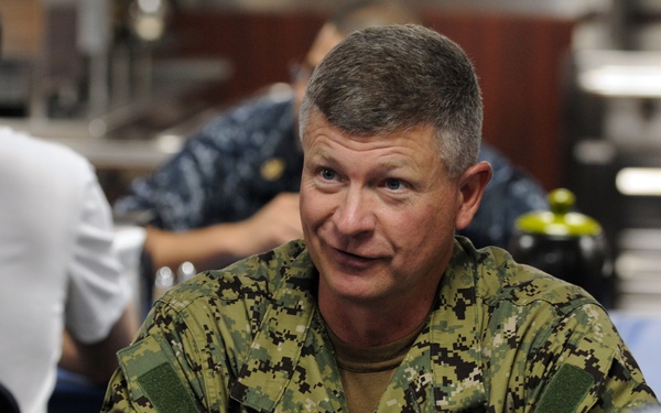 A senior military officer wearing US Navy Working Uniform (NWU) Type III (woodland camouflage pattern) is seated at a table engaged in conversation with other personnel. The officer's name tape partially reads 'NEST' and branch tape appears to read 'U.S. N[AVY]'. The setting appears to be a military dining facility or mess hall.