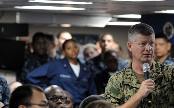 A senior U.S. Navy officer in NWU (Navy Working Uniform) camouflage is addressing a large group of sailors in a shipboard compartment. He is holding a microphone and gesturing, suggesting a command briefing or all-hands meeting. A blue banner with the word 'BLUE' is visible in the background.