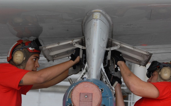Two military personnel in red shirts and camouflage pants are working on an aircraft weapons pylon in what appears to be a naval vessel hangar bay. They are handling a blue-tipped ordnance or training munition being mounted or secured to the underside of an aircraft wing. Both individuals are wearing red cranials (helmets) with hearing protection, indicating standard naval aviation safety gear.