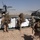 Supporting, Transporting Troops: HMH-361 Flies Across Helmand Province Military personnel in full combat gear with large backpacks are gathered on an arid airfield alongside white cargo containers. Two large military helicopters, identifiable as CH-53 Sea Stallions, are on or near the ground with rotors spinning. A third helicopter is visible in the distant background, suggesting an active air assault or logistics operation in a desert environment.