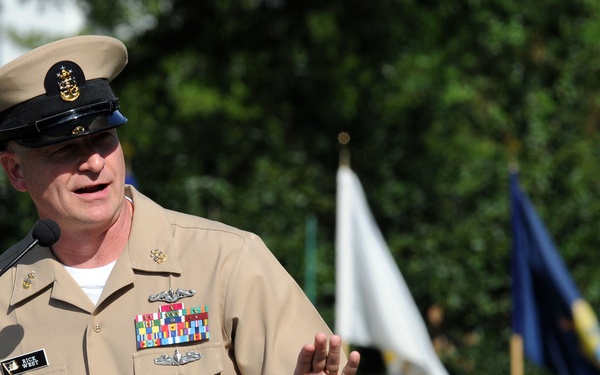 A U.S. Navy senior enlisted officer, identifiable by khaki service uniform and combination cover with anchor insignia, is speaking at a podium with a microphone at an outdoor military ceremony. He is gesturing with his right hand while addressing an audience. Multiple flags, including what appears to be a white ceremonial flag and blue Navy flags, are visible in the background.