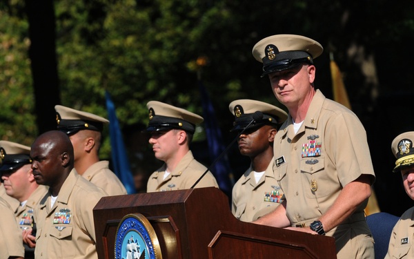 A formal U.S. Navy ceremony is taking place outdoors, with a senior enlisted sailor speaking at a podium bearing the official seal of the Department of the Navy. Multiple Navy personnel in Service Khaki (Type B) uniforms are standing in formation, while a senior officer with gold-braided cap sits nearby. An American flag is draped at the base of the podium, and additional flags are visible in the background.