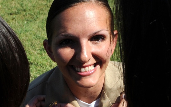 A smiling woman in a tan/khaki military uniform is having insignia or rank pins attached to her collar by two other individuals. The scene appears to be a military promotion or commissioning ceremony taking place outdoors on a grassy area. The central figure appears joyful and proud during what is likely a significant milestone event.