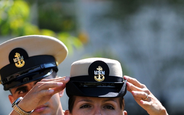 A U.S. Navy female Chief Petty Officer is having her combination cover (cap) placed on her head during what appears to be a Chief Petty Officer pinning or advancement ceremony. A male Navy Chief Petty Officer stands behind her, placing the cover on her head. Both are wearing the Navy Service Uniform (NSU) in khaki/tan.