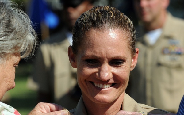 A military promotion or pinning ceremony is taking place outdoors. A smiling female service member in a tan/khaki military uniform is having insignia or rank devices pinned to her collar by two civilians, one on each side. The background shows other uniformed military personnel, suggesting an official ceremony.
