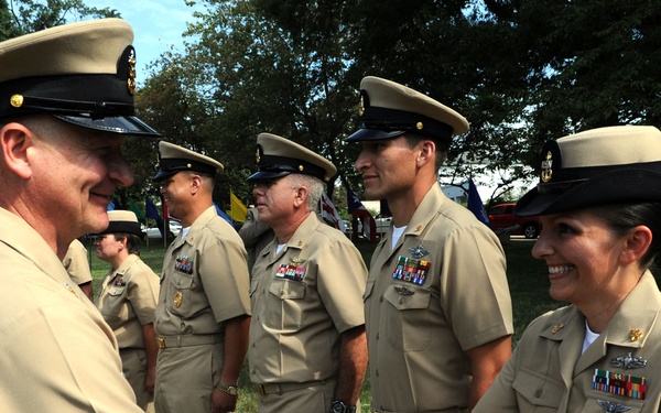 A group of U.S. Navy personnel in tan/khaki service uniforms are gathered outdoors on a grassy area, likely during a formal military ceremony or event. A male sailor on the left is shaking hands with a female sailor on the right, both smiling. Several other uniformed personnel stand in the background observing.