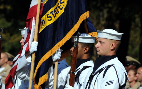 A United States Navy color guard is performing a ceremonial parade or formal military ceremony. Sailors in dress white uniforms carry the American flag and a Navy unit flag with gold fringe, while also bearing ceremonial rifles. In the background, additional Navy personnel in khaki/tan service uniforms stand in formation as observers.