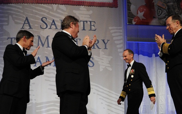 A formal military ceremony or gala event is taking place on a stage, with a naval officer in dress uniform walking toward applauding individuals in black tuxedos. A banner in the background reads 'A Salute to Military Families.' The scene suggests an award presentation or recognition ceremony honoring military service.