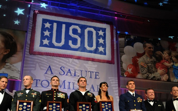 A formal USO awards ceremony titled 'A Salute to the Military' featuring a group of military personnel and civilians standing on stage holding framed award plaques. The backdrop displays the USO logo along with projected images of military families. The event appears to be a gala or formal recognition ceremony honoring service members.