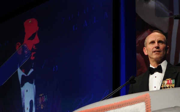 A military officer in formal dress uniform with medals is speaking at a podium bearing the USO logo at what appears to be the 2010 USO Gala. He is wearing a black tuxedo with military decorations on the left chest. A large projection screen behind him displays his image alongside event branding.