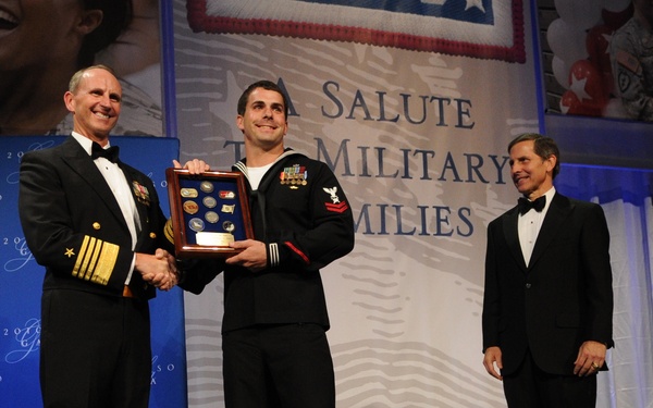 A formal military awards ceremony is taking place on a stage, where a high-ranking Navy admiral in dress uniform is presenting a shadow box display case containing coins and medals to a Navy enlisted sailor in dress blues. The backdrop reads 'A Salute to Military Families,' suggesting this is a USO or similar military appreciation event. Two civilians in tuxedos stand nearby as witnesses to the presentation.