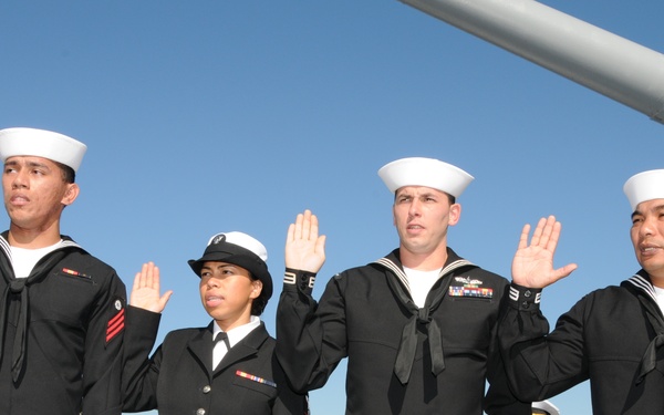 Four U.S. Navy personnel are raising their right hands in what appears to be a reenlistment or enlistment ceremony conducted on the deck of a naval vessel. Three sailors are wearing the Navy's dress blue 'Crackerjack' uniform with white Dixie cup covers, while one female sailor wears the Service Dress Blue coat uniform with a combination cover. A large naval gun barrel is visible in the upper right background.