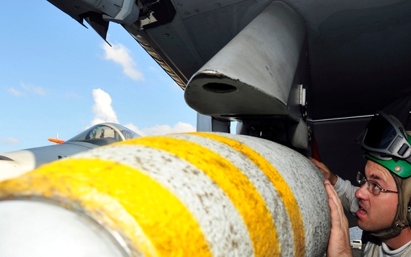 A military ordnance technician wearing a flight deck helmet and hearing protection is inspecting or loading a large bomb/munition onto the underside of a military aircraft. The munition features distinctive yellow bands, which are visible markings used to indicate the type or status of the ordnance. The setting appears to be the flight deck of an aircraft carrier, with another jet aircraft visible in the background.