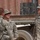 Three U.S. Army soldiers in ACU (Army Combat Uniform) digital camouflage are engaged in conversation near a military vehicle and shipping container. Two soldiers face each other in discussion while a third stands nearby. The setting appears to be a forward operating base or logistics area.
