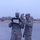 Two military personnel in US Army ACU (Army Combat Uniform) digital camouflage are standing outdoors on a gravel surface in low-light or hazy conditions. One soldier is holding a black tactical device with an antenna and a document or label attached to it, while the second soldier appears to be pointing at or adjusting the device. The background shows sparse trees and an overcast or dusty sky.