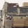 A U.S. Army soldier in Army Combat Uniform (ACU) digital camouflage pattern stands facing a tan/desert-colored armored military vehicle, holding what appears to be a portable electronic device or control unit up toward the vehicle. The vehicle appears to be a Mine-Resistant Ambush Protected (MRAP) or similar heavy armored wheeled vehicle equipped with multiple electronic systems and antennas. The setting appears to be a gravel-covered military base or staging area in an arid environment.