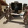 U.S. Army soldiers in ACU (Army Combat Uniform) are carrying a person on a litter/stretcher away from a military vehicle, likely a HMMWV (Humvee). A second individual is visible stepping out of the rear of the vehicle. The scene appears to take place at a forward operating base or military compound in an arid environment.