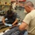 Two medical personnel wearing blue latex gloves are attending to a patient lying on a Stryker gurney in what appears to be a military or emergency medical facility. One individual is wearing dark gray scrubs and the other is in a tan/khaki t-shirt, both focused on the patient's lower extremities. An American flag is partially visible in the background, and the room is stocked with medical equipment and supplies.