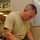 A male individual wearing U.S. Army ACU (Army Combat Uniform) digital camouflage pants and an olive drab t-shirt is writing on paperwork placed on a metal tray stand. He is wearing blue nitrile examination gloves and has what appears to be a holstered sidearm on his left hip. The setting appears to be a medical or clinical facility based on the institutional interior visible in the background.