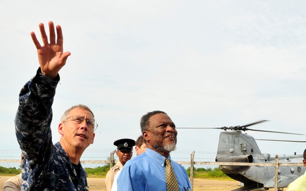 A group of individuals, including a person in U.S. Navy digital camouflage uniform, are walking across an open dirt field. The uniformed individual is waving with one raised hand while walking alongside civilians. A military tandem-rotor helicopter, consistent with a CH-46 Sea Knight, is visible in the background behind a chain-link fence.