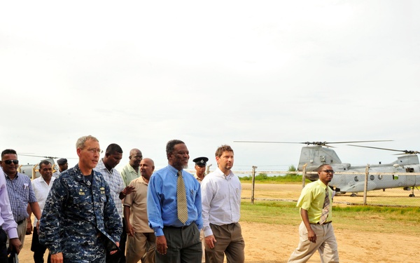 A group of approximately 10-12 individuals, including one person in U.S. Navy digital camouflage (NWU) uniform and several civilians in business casual attire, are walking across a dirt field. In the background, two military helicopters identified as CH-46 Sea Knight tandem-rotor helicopters are parked behind a chain-link fence. The setting appears to be a temporary or forward operating airfield in a tropical or coastal environment.