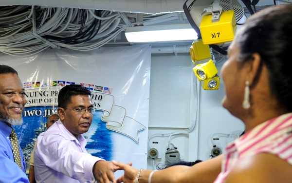 The image shows a social gathering aboard what appears to be a naval vessel, likely the USS Iwo Jima, during a 2010 event. Three individuals are prominently featured in a handshaking or greeting interaction, with a backdrop banner partially visible referencing 'Iwo Jima' and '2010'. The setting appears to be an interior deck space with industrial lighting and cable management systems overhead.