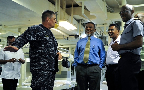 A U.S. Navy sailor in digital camouflage uniform appears to be conducting a tour or briefing for a group of civilian visitors inside what appears to be a berthing compartment aboard a naval vessel. The sailor is gesturing with an outstretched arm, seemingly explaining the space to the attentive group. The civilians are dressed in business casual attire and appear engaged and smiling.