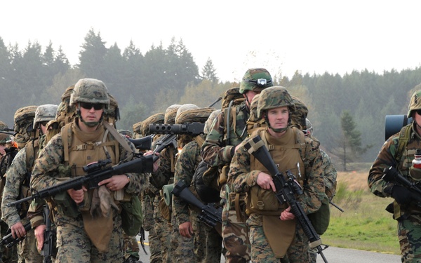 A large column of military personnel conducting a road march along a paved road in what appears to be a training environment. The soldiers are fully equipped with combat gear, rucksacks, helmets, and weapons. The setting appears to be a military installation surrounded by coniferous trees and open fields.