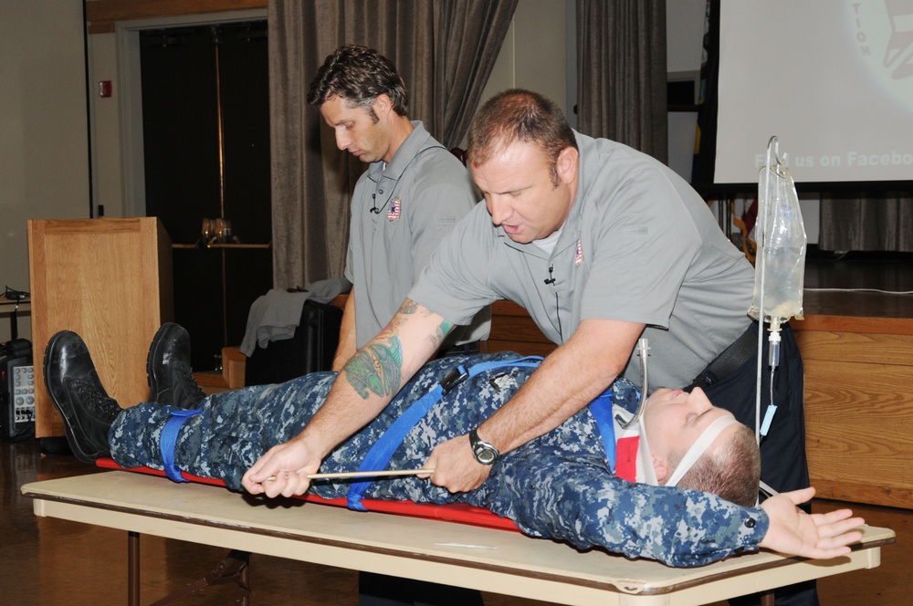 Two individuals in gray uniforms are conducting what appears to be a medical training demonstration. A person in Navy digital camouflage (NWU) uniform is lying on a table secured to a backboard with straps and a cervical collar, while one instructor leans over to demonstrate a procedure. An IV bag is visible on a stand nearby, suggesting this is a first aid or combat casualty care training session.