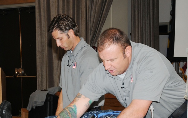 Two individuals in gray uniforms are conducting what appears to be a medical training demonstration. A person in Navy digital camouflage (NWU) uniform is lying on a table secured to a backboard with straps and a cervical collar, while one instructor leans over to demonstrate a procedure. An IV bag is visible on a stand nearby, suggesting this is a first aid or combat casualty care training session.