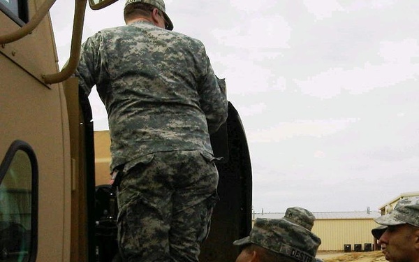 Several U.S. Army soldiers in ACU (Army Combat Uniform) digital camouflage pattern are gathered around a tan/beige military vehicle, likely a HMMWV (Humvee). One soldier is climbing into or inspecting the vehicle while others observe from ground level. The setting appears to be a military installation or training area with a building visible in the background.
