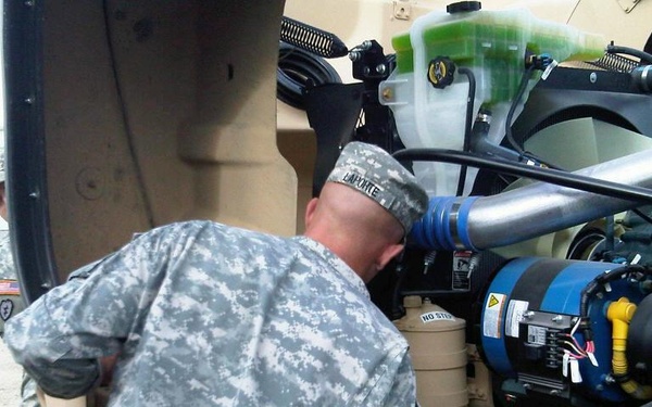 Two U.S. Army soldiers in ACU (Army Combat Uniform) are performing maintenance or inspection on a military vehicle engine compartment. The primary soldier, identified by name tape 'LAPORTE' on his patrol cap, is leaning into the engine bay examining components. The setting appears to be a military installation or motor pool area.