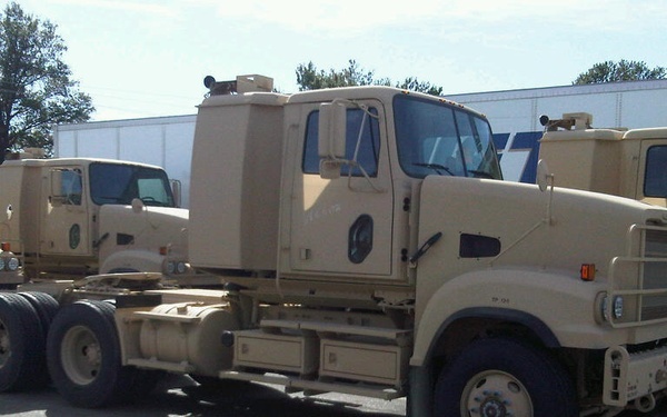 The image shows multiple military-style semi-truck tractors (cab-over and conventional configurations) painted in desert tan/sand color, parked in what appears to be a commercial parking lot or truck stop. The vehicles appear to be surplus military trucks, possibly being staged or transported. A commercial building and fuel price sign are visible in the background.