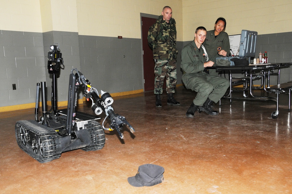 Military personnel are conducting a demonstration or training exercise with a tracked EOD (Explosive Ordnance Disposal) robot inside what appears to be a military facility or cafeteria. One operator is seated at a control station with a laptop, while two other personnel observe. The robot appears to be approaching or examining a military cap placed on the floor.