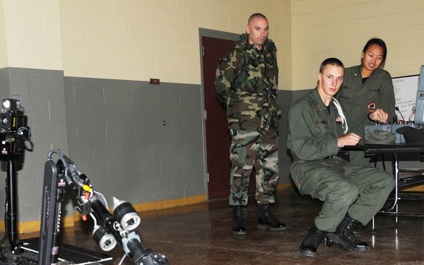 Military personnel are conducting a demonstration or training exercise with a tracked EOD (Explosive Ordnance Disposal) robot inside what appears to be a military facility or cafeteria. One operator is seated at a control station with a laptop, while two other personnel observe. The robot appears to be approaching or examining a military cap placed on the floor.