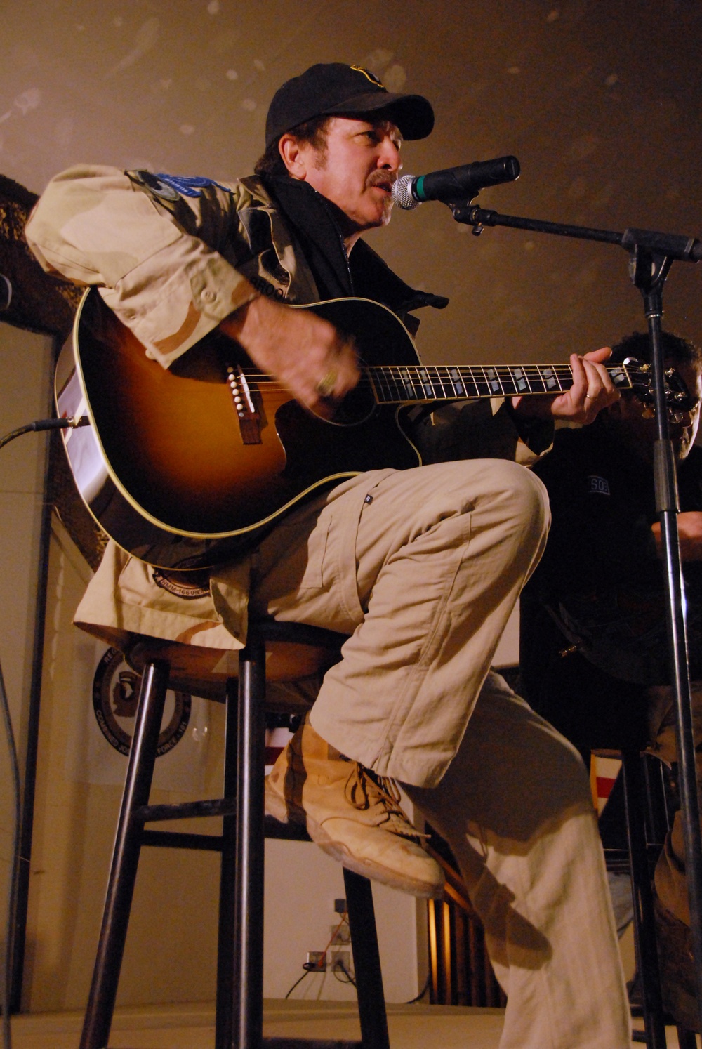 A middle-aged man is performing live music, seated on a bar stool while playing an acoustic guitar and singing into a microphone. He is wearing a black baseball cap, a camouflage jacket over a black shirt, khaki cargo pants, and tan lace-up boots. A second person is partially visible in the background to the right.