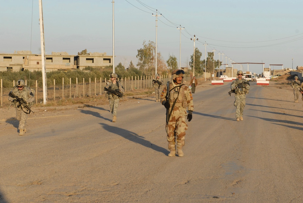 The image shows a joint military patrol on a road in what appears to be a Middle Eastern urban or semi-urban environment. Multiple soldiers in different uniforms — including US Army ACU (Army Combat Uniform) pattern and a desert tri-color camouflage pattern worn by what appears to be an Iraqi Security Forces soldier — are walking along a road while carrying weapons. The setting features utility poles, concrete block buildings, a perimeter fence, and a checkpoint structure in the background.