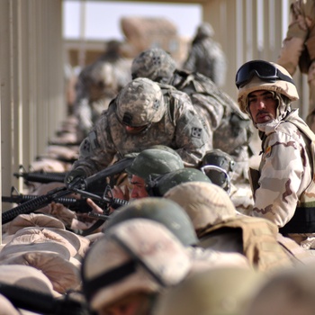 The image shows a military training or firing range exercise with soldiers in prone shooting positions along a covered range lane. Two distinct uniform types are visible: US Army ACU (Army Combat Uniform) pattern and a desert tri-color camouflage pattern, suggesting a joint training exercise. Soldiers are positioned along what appears to be a covered outdoor firing range in an arid environment.