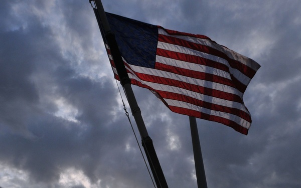 A dramatic low-angle photograph captures an American flag flying from a mast against a stormy, overcast sky at what appears to be dusk or dawn. Several silhouetted figures in military-style uniforms stand on what appears to be the deck of a naval vessel or military installation. The backlighting from the partially obscured sun creates a high-contrast, atmospheric composition.