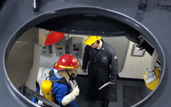 The image is taken from above through a circular hatch or opening on what appears to be a naval vessel, looking down into an interior compartment. Two personnel are visible below: one wearing a red hard hat, blue coveralls, a face shield, and white gloves, and another in dark coveralls with a yellow hard hat and headset holding a clipboard or document. The setting and equipment suggest a safety inspection or damage control drill aboard a military ship.